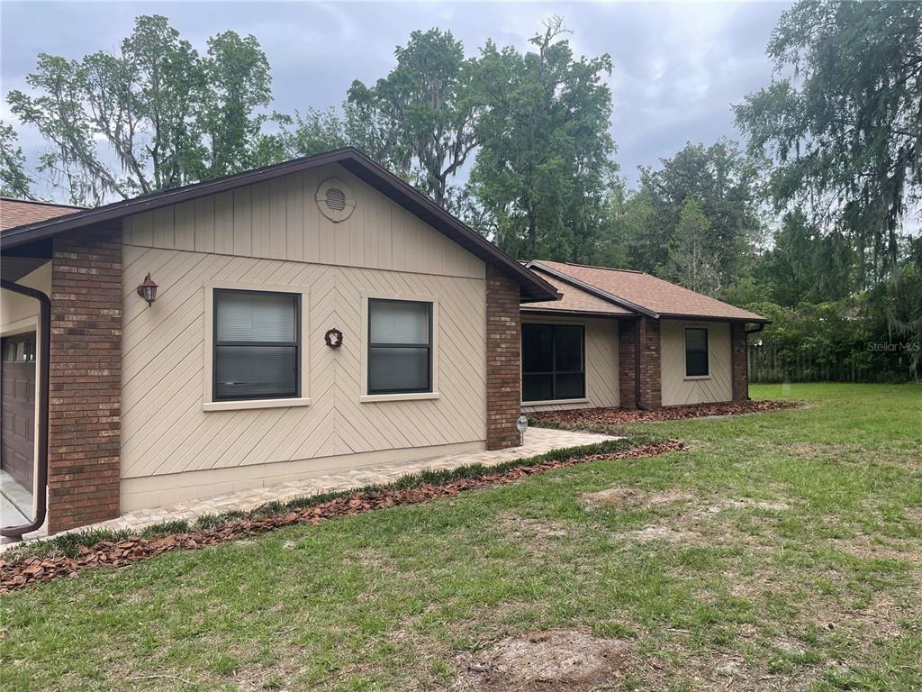 A single-story house with tan siding and brick accents sits on a grassy, wooded lot — 2421 NW 106TH DRIVE, GAINESVILLE, FL