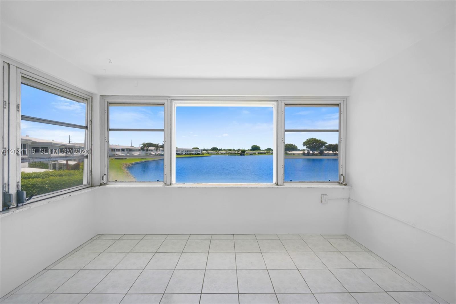 A bright, tiled room features large windows overlooking a lake and surrounding residential buildings — 1116 Lake Ter # 208, Boynton Beach FL 33426, BOYNTON BEACH, FL