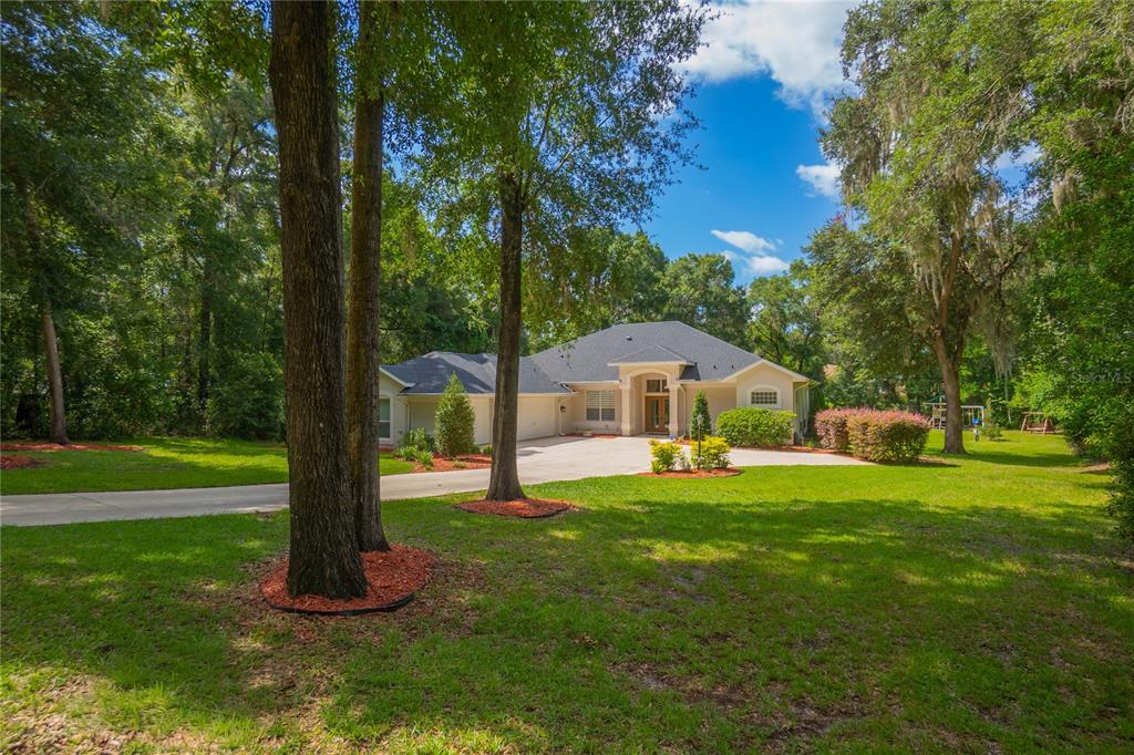 A single-story house with a concrete driveway sits on a wooded lot under a blue sky — 18976 SW 93RD LOOP, DUNNELLON, FL