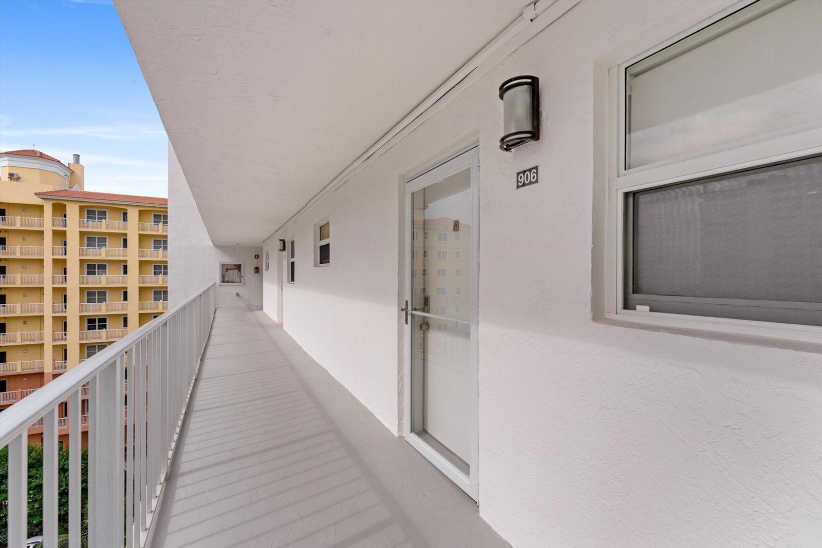 An exterior walkway features white walls, a numbered door, and a metal railing overlooking buildings — 1391 S Ocean Boulevard S # 906, Pompano Beach FL 33062, POMPANO BEACH, FL