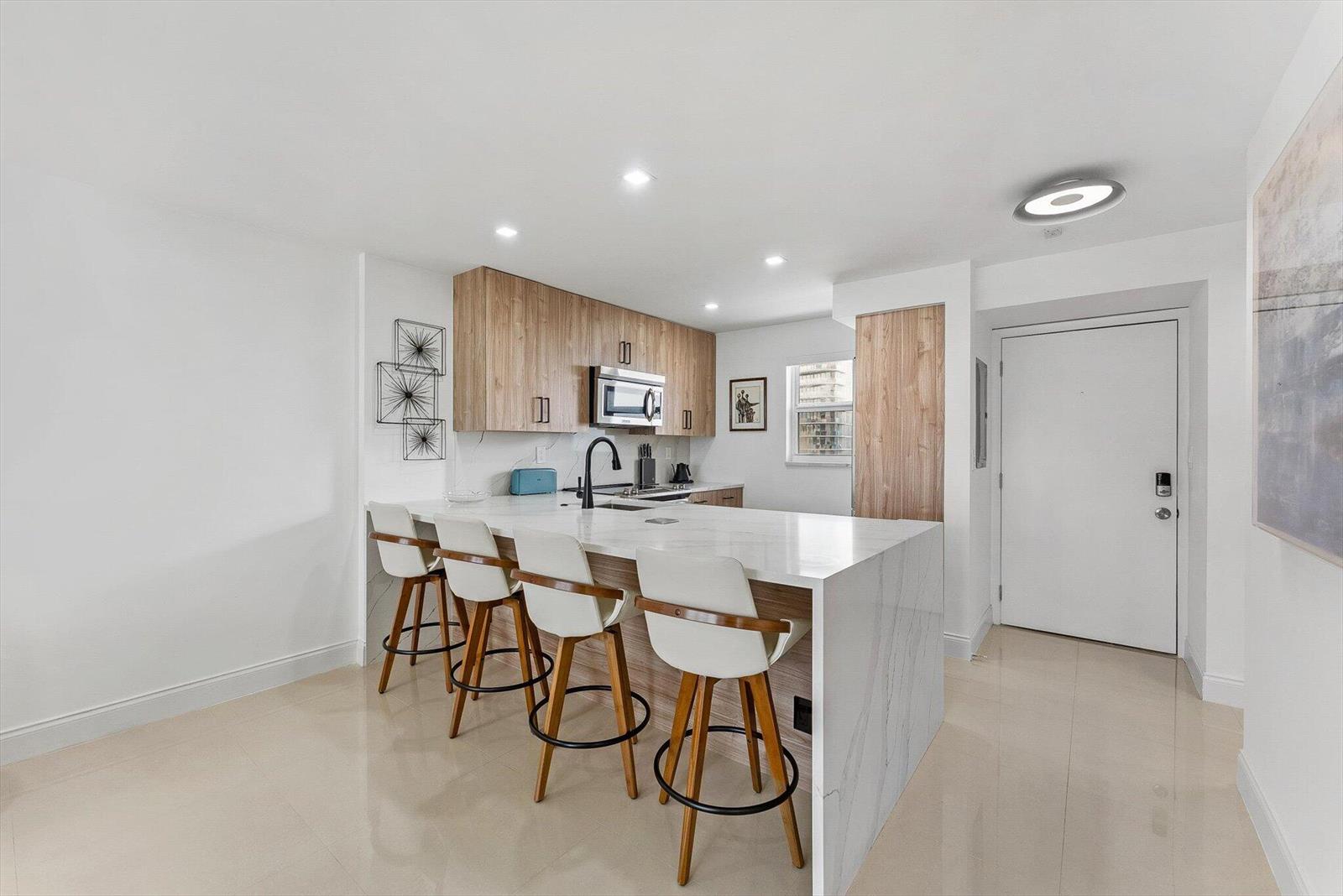A modern kitchen features a marble waterfall island with four bar stools and wood cabinetry — 1391 S Ocean Boulevard S # 906, Pompano Beach FL 33062, POMPANO BEACH, FL