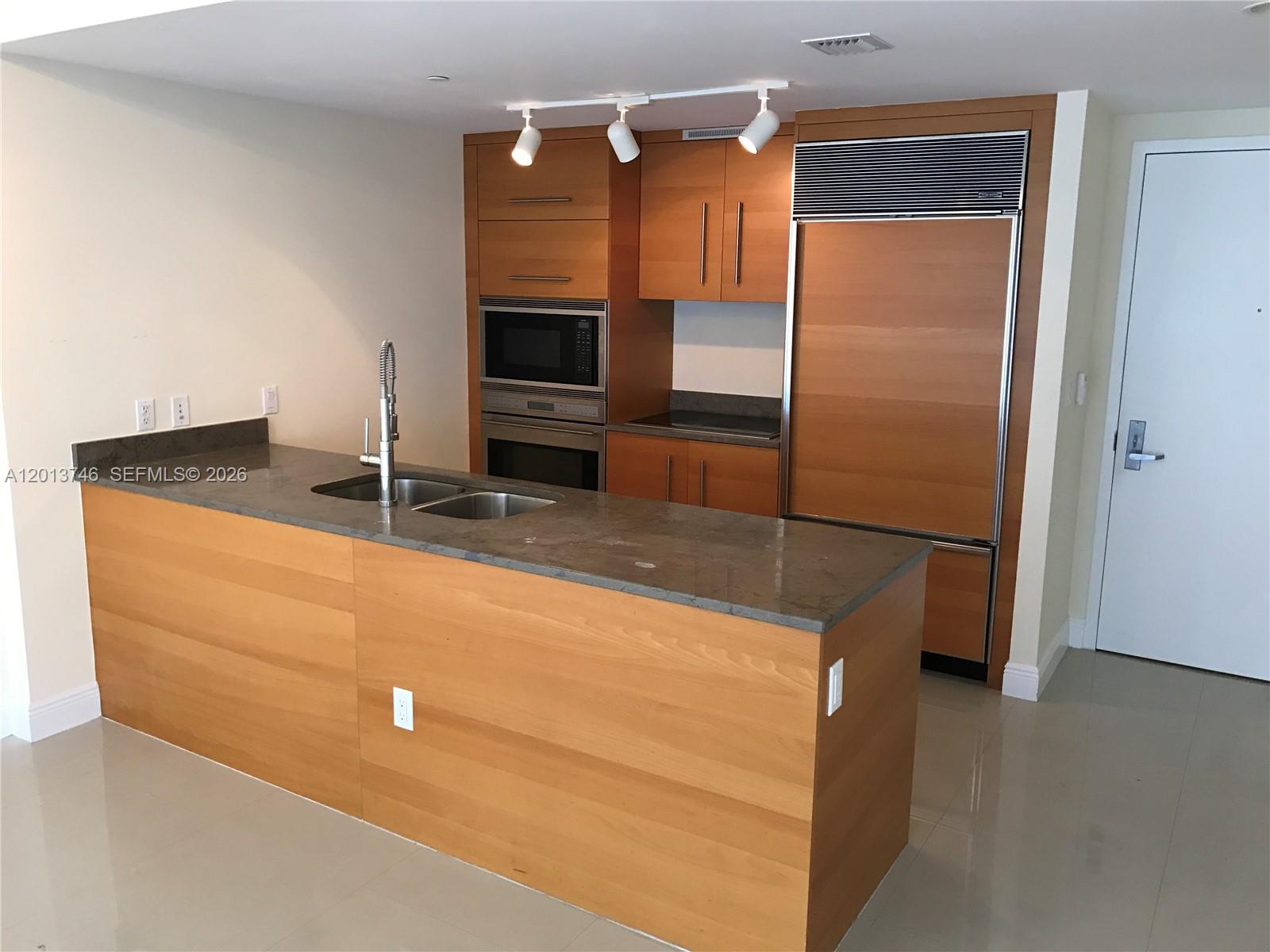 A kitchen features wood cabinetry, a gray stone countertop, and a stainless steel refrigerator — 475 Brickell Ave # 2411, Miami FL 33131, MIAMI, FL