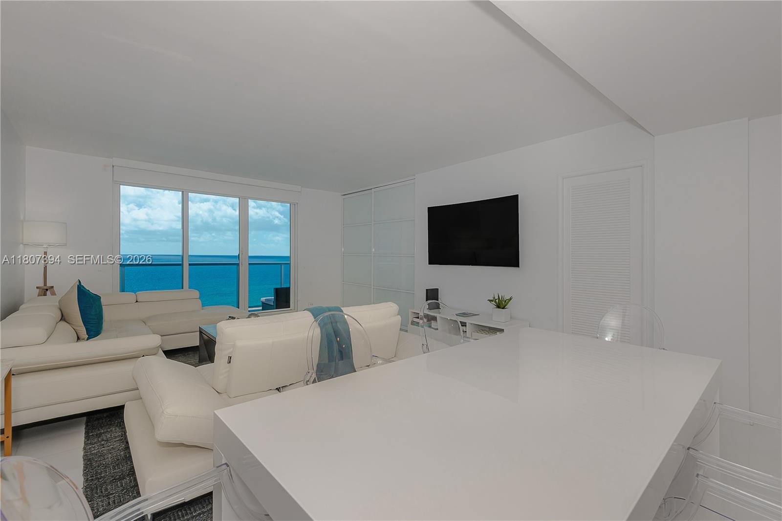 A modern living room features white furniture, a wall-mounted television, and floor-to-ceiling ocean views — 2301 Collins Ave # 1538, Miami Beach FL 33139, MIAMI BEACH, FL