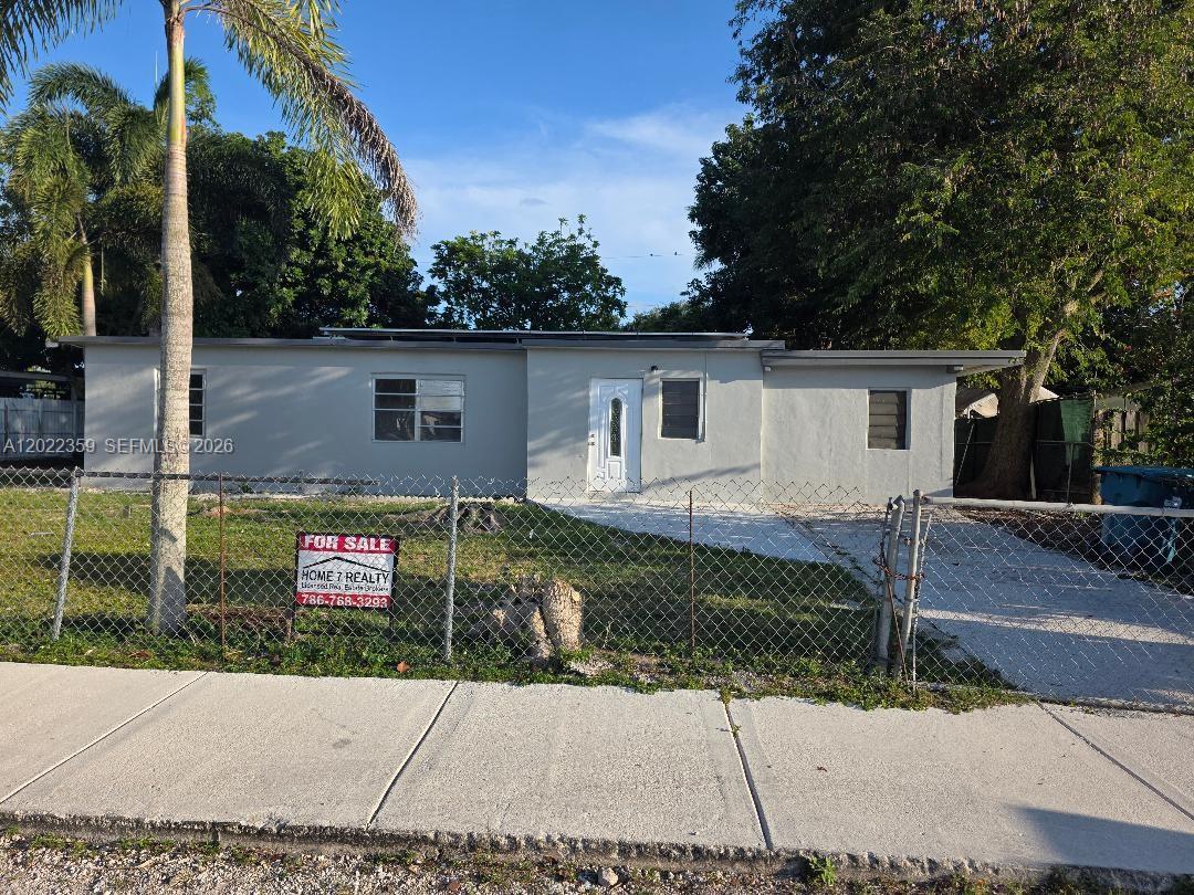 A single-story gray house with a white door sits behind a chain-link fence — HOMESTEAD, FL