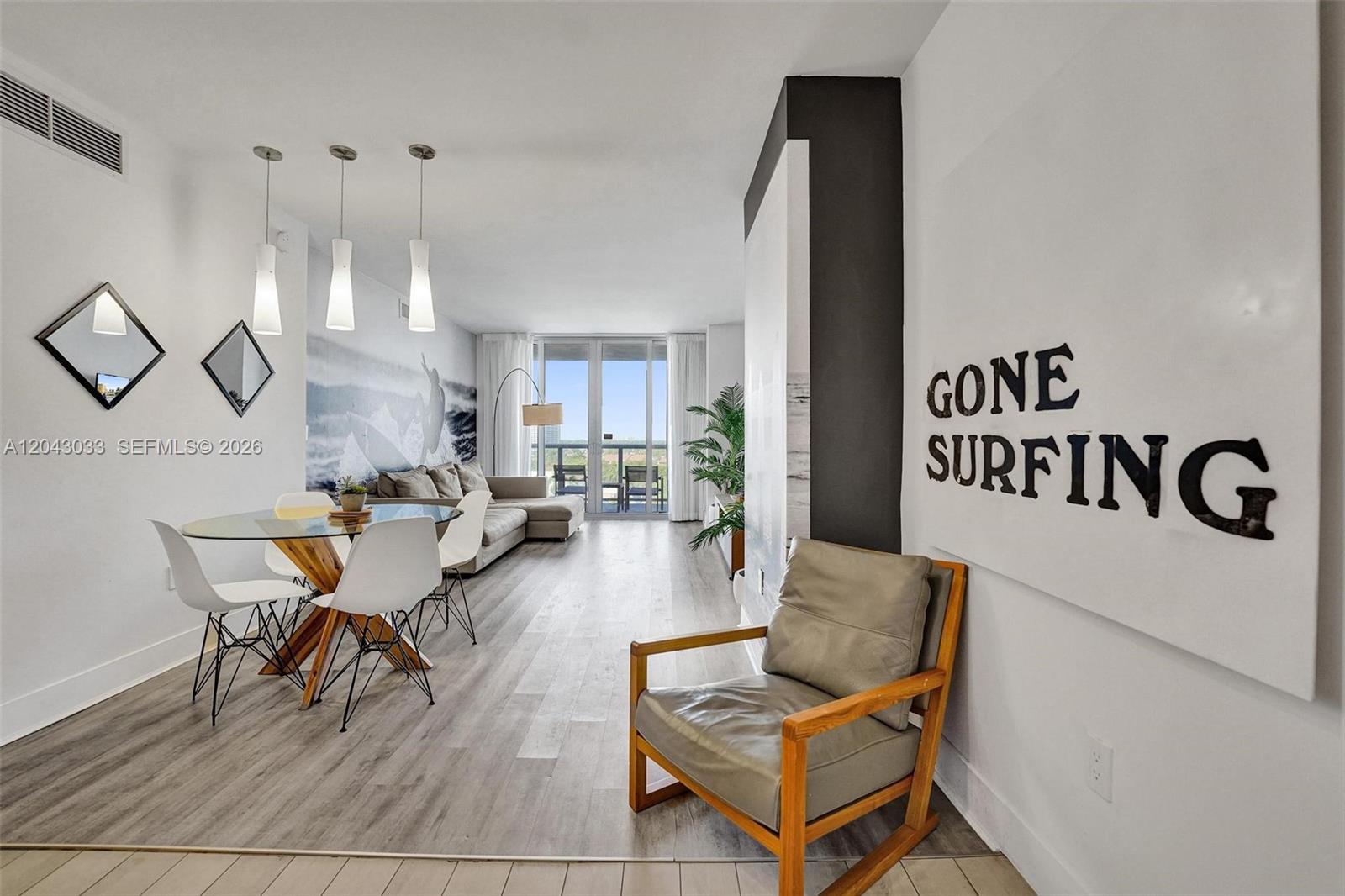 A modern living and dining area features light wood floors, a glass table, and surf-themed decor — 2602 E Hallandale Beach Blvd # R906, Hallandale Beach FL 33009, HALLANDALE BEACH, FL