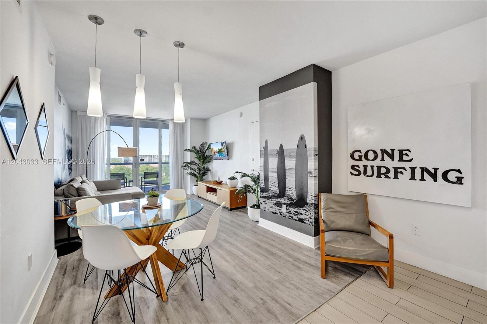 A modern living and dining area features a glass table, surf-themed decor, and balcony access — 2602 E Hallandale Beach Blvd # R906, Hallandale Beach FL 33009, HALLANDALE BEACH, FL