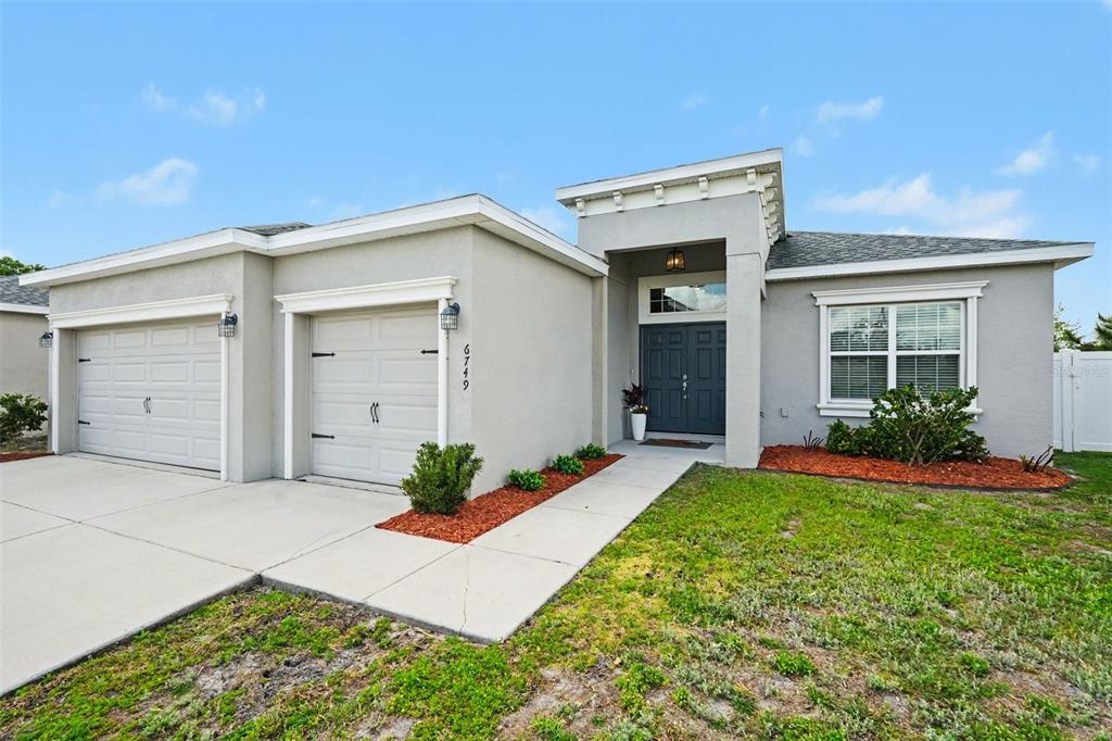 A single-story house with a two-car garage, gray stucco exterior, and a dark front door — 6749 ABBYWOOD LANE, ZEPHYRHILLS, FL