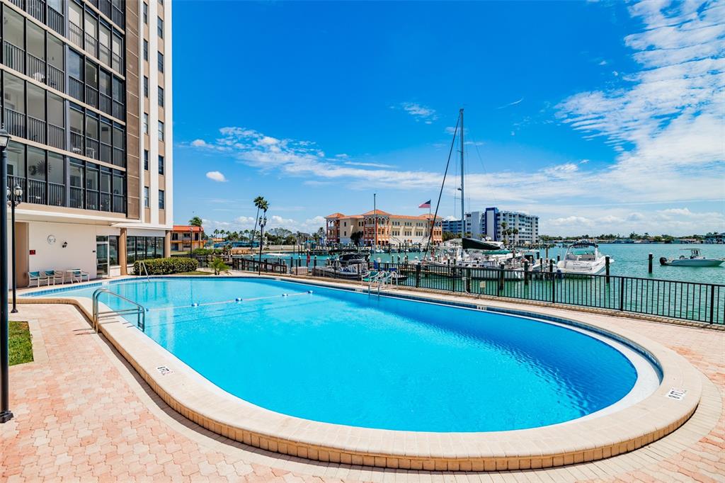 A swimming pool sits on a paved deck next to a high-rise building and marina — 10355 PARADISE BOULEVARD UNIT 511, TREASURE ISLAND, FL