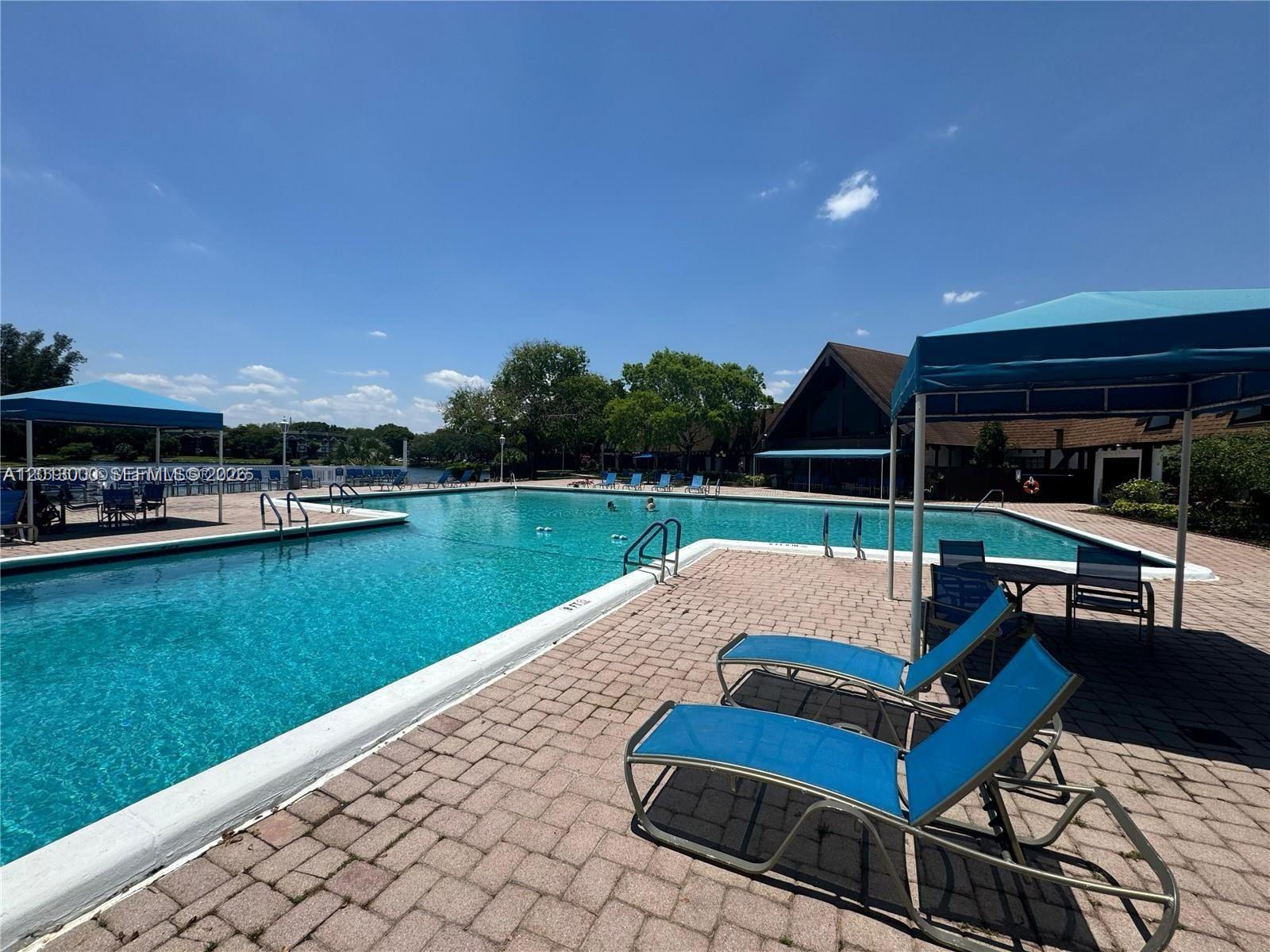 A large outdoor swimming pool with lounge chairs and shaded seating areas on a brick deck — 3730 Inverrary Dr # 3B, Lauderhill FL 33319, LAUDERHILL, FL