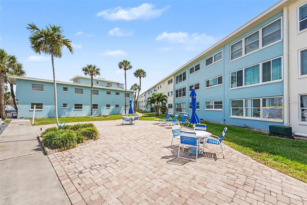 A paved patio area with outdoor tables and chairs sits between two light blue apartment buildings — 6161 GULF WINDS DRIVE UNIT 153, ST PETE BEACH, FL