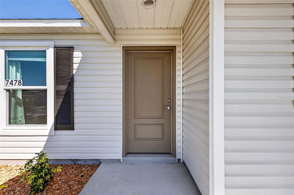 The exterior of a house features a tan front door, white siding, and a window — 7478 SAVANNA DRIVE, THE VILLAGES, FL