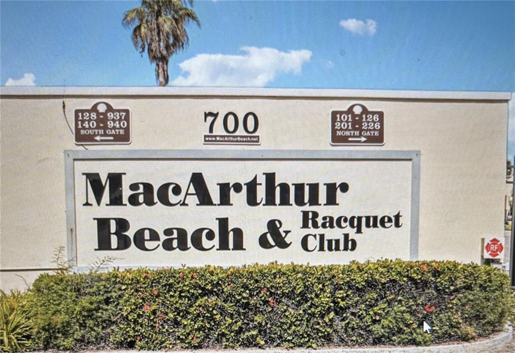 A sign for MacArthur Beach & Racquet Club stands above a hedge with directional gate information — 700 GOLDEN BEACH BOULEVARD UNIT 130, VENICE, FL