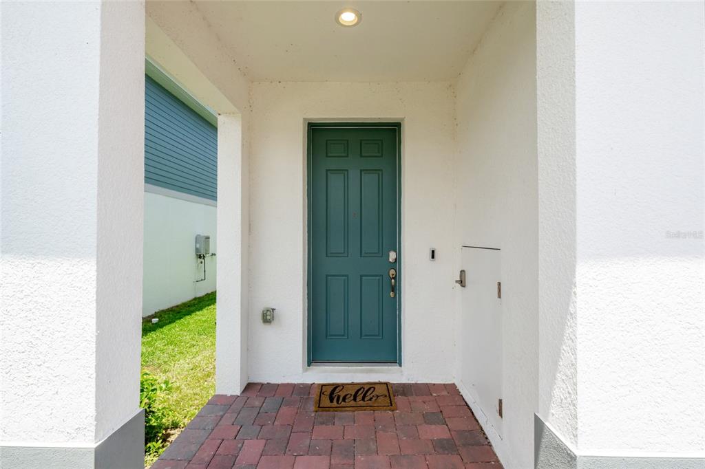 A teal front door with a welcome mat sits under a covered entryway with pavers — 6809 SERPENT HAWK LANE, HOWEY IN THE HILLS, FL