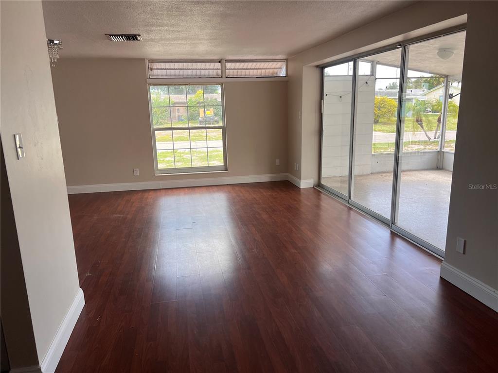 An empty living room features dark wood-look flooring, a large window, and sliding glass doors — 6111 ROLLINS STREET, BRADENTON, FL