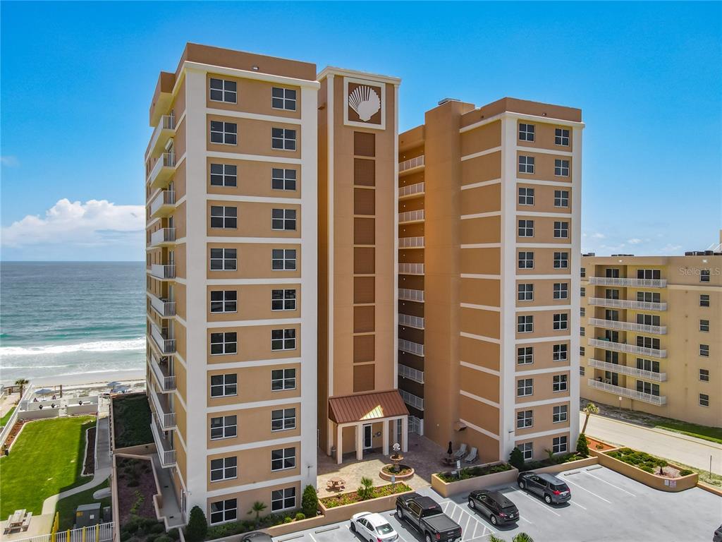 An aerial view of a tan, multi-story beachfront condominium building with a shell logo — 3799 S ATLANTIC AVENUE UNIT 1001, DAYTONA BEACH, FL