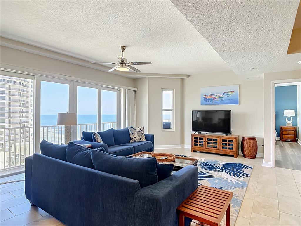 A living room features a blue sofa, a television, and a balcony with ocean views — 3799 S ATLANTIC AVENUE UNIT 1001, DAYTONA BEACH, FL