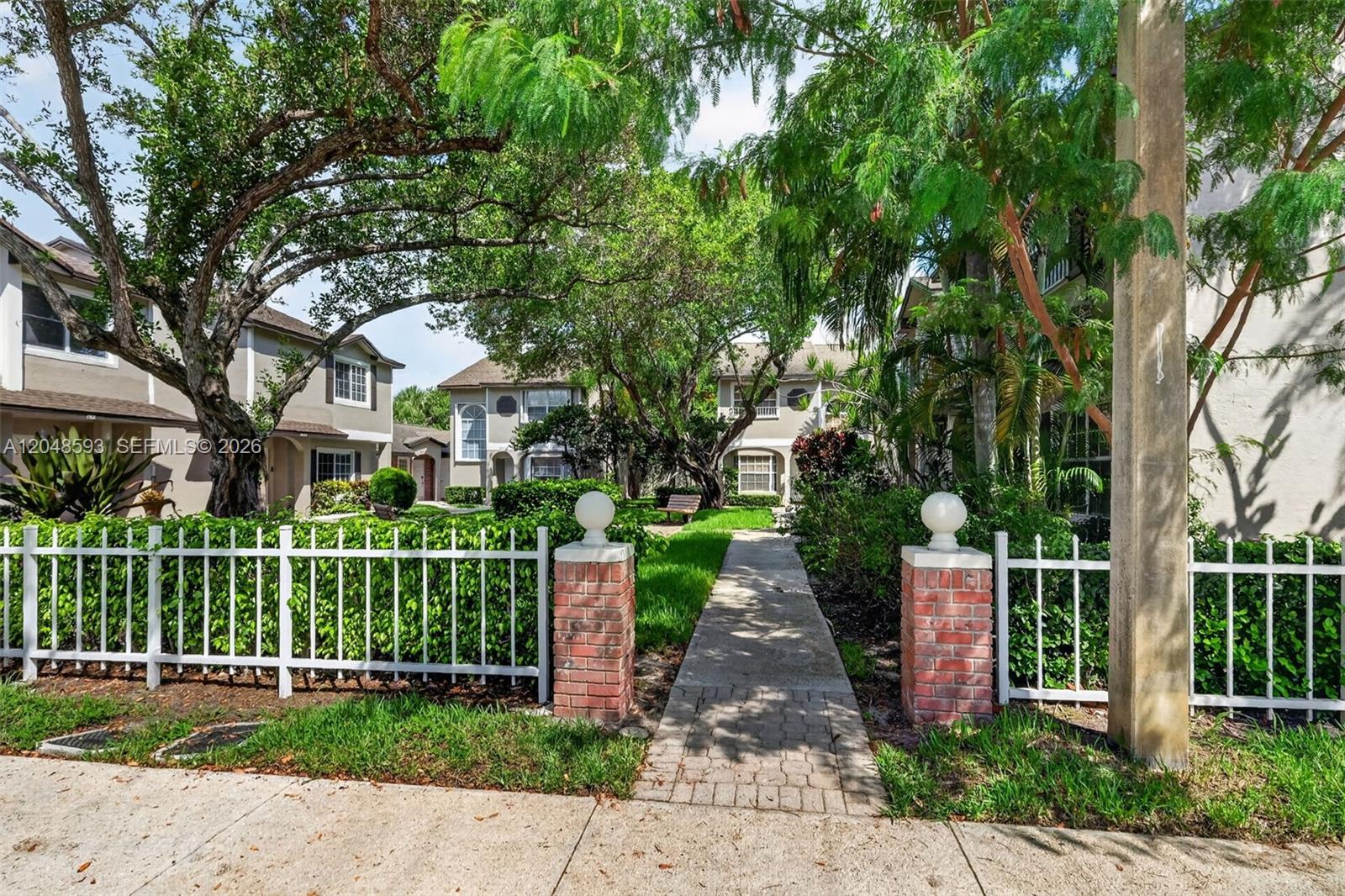 A paved walkway leads through a brick-pillared gate toward residential buildings surrounded by mature trees — 3835 NW 122nd Ter, Sunrise FL 33323, SUNRISE, FL
