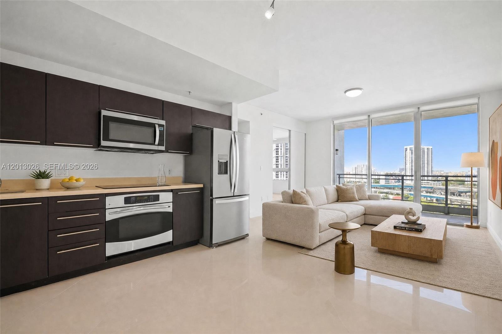 A modern kitchen with dark cabinetry and stainless steel appliances opens into a bright living area — 90 SW 3rd St # 1608, Miami FL 33130, MIAMI, FL
