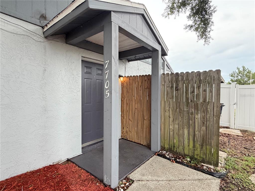 The exterior view shows a covered entryway with a gray door and a wooden fence — 7705 CITRONELLA COURT, TAMPA, FL