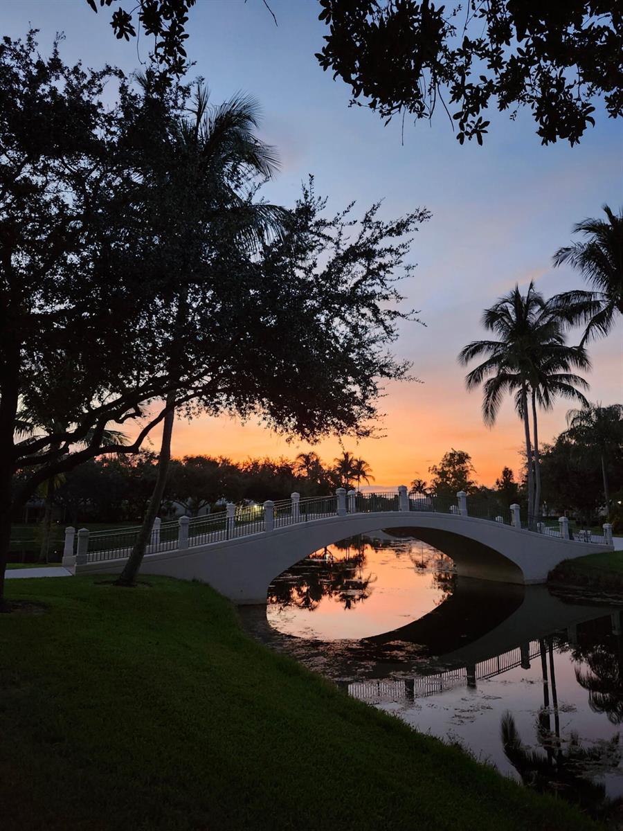 An arched pedestrian bridge crosses a calm canal at sunset, framed by palm trees and foliage — 8093 Montserrat Place, Wellington FL 33414, WELLINGTON, FL