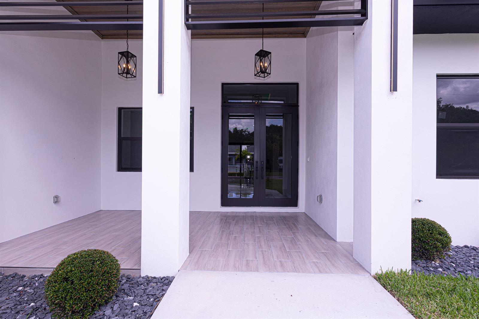 A modern home entrance features a covered porch with wood-look tile and double glass doors — 6574 Rigger Road, Lake Worth FL 33462, LAKE WORTH, FL