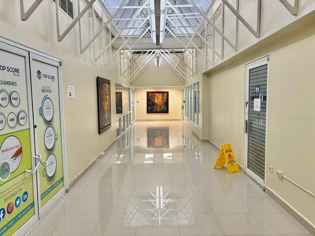 A bright, tiled hallway features a glass-paneled ceiling, framed artwork, and a yellow caution sign — 300 AVENIDA LAS CUMBRES, SAN JUAN, PR