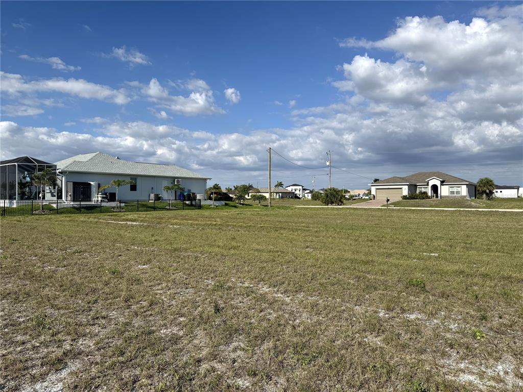 A large, grassy vacant lot sits between two suburban houses under a blue sky — 3436 NW 45TH AVENUE, CAPE CORAL, FL