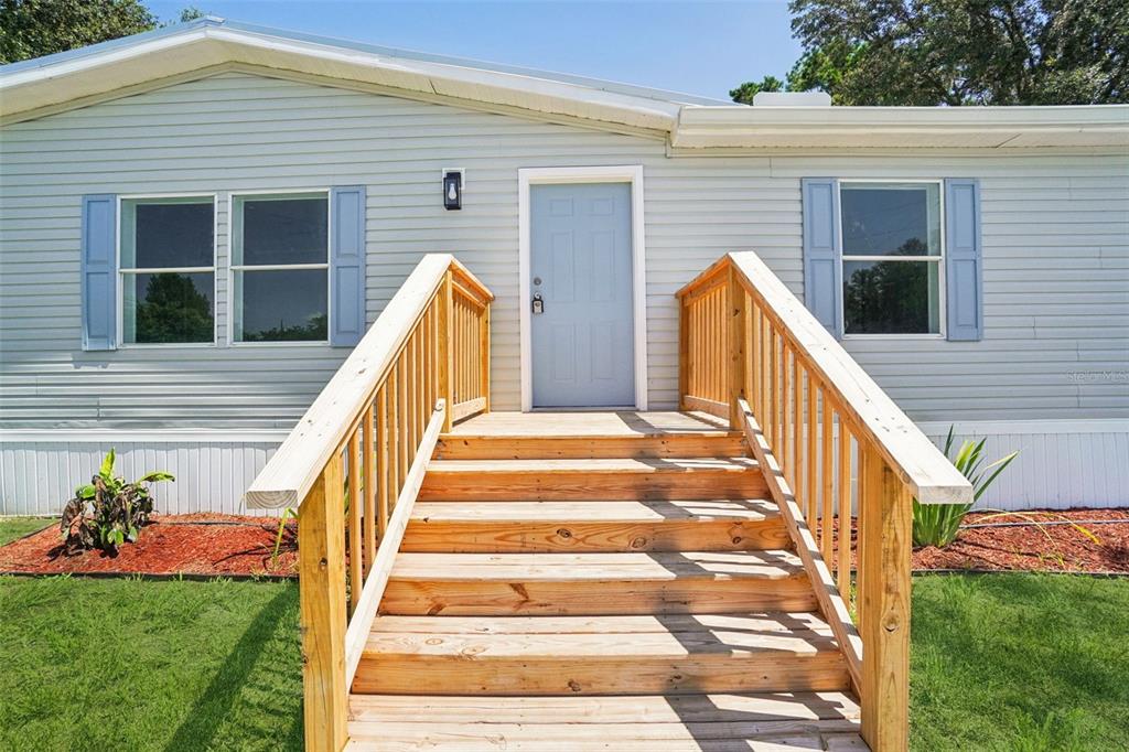 A light gray mobile home exterior features a wooden staircase leading to a blue front door — 29417 KELLY DRIVE, WESLEY CHAPEL, FL