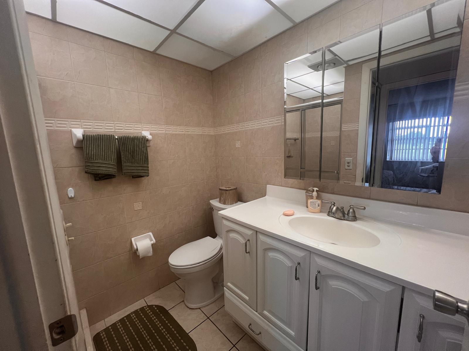 A bathroom features beige tiled walls, a white vanity, a toilet, and a medicine cabinet — A Flanders # 5, Delray Beach FL 33484, DELRAY BEACH, FL