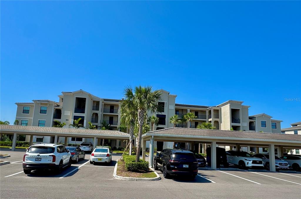 A multi-story apartment building with covered parking spaces and several parked cars in the foreground — 18028 GAWTHROP DRIVE UNIT 303, BRADENTON, FL