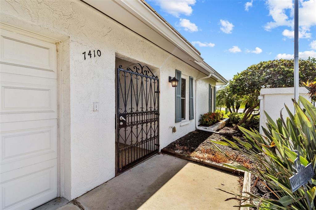 A white stucco house exterior features a black metal gate, a window with shutters, and landscaping — 7410 8TH AVENUE W, BRADENTON, FL