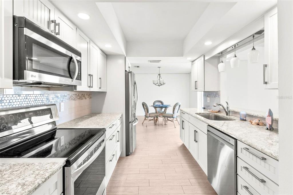A galley kitchen with white cabinets, stainless steel appliances, and light countertops leads to a dining area — 7410 8TH AVENUE W, BRADENTON, FL