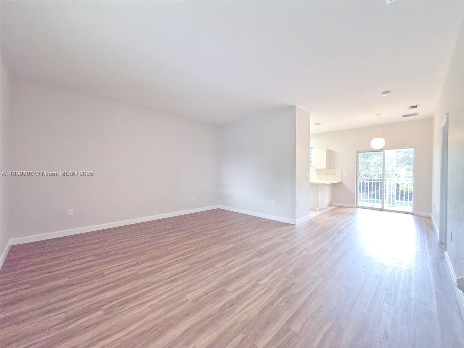An empty living area with light wood-look flooring leads to a kitchen and balcony — 2610 NE 3rd Dr # 203, Homestead FL 33033, HOMESTEAD, FL