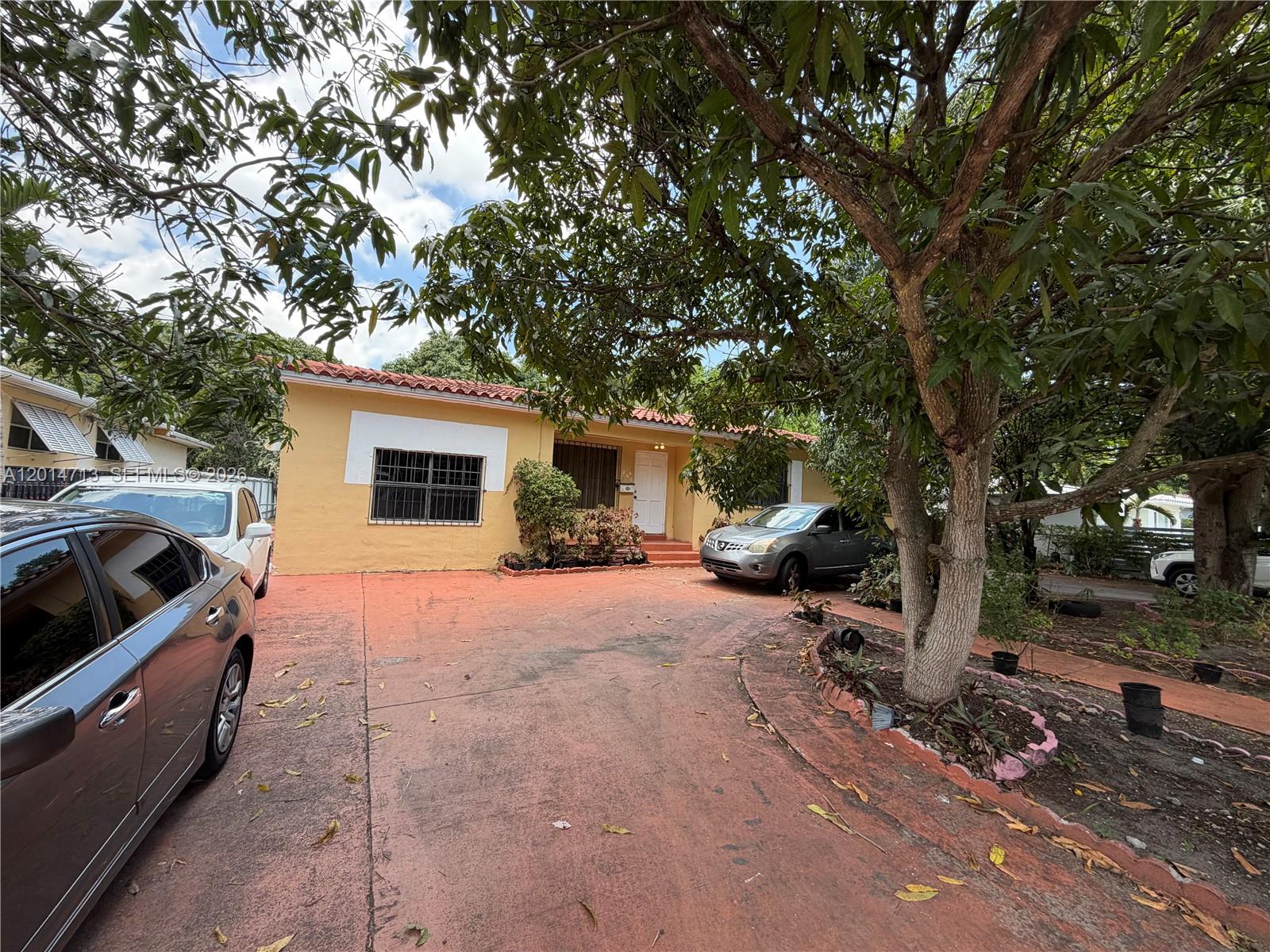 A yellow single-story house with a red-tiled roof sits behind a large tree and driveway — 385 NE 129th St, North Miami FL 33161, NORTH MIAMI, FL