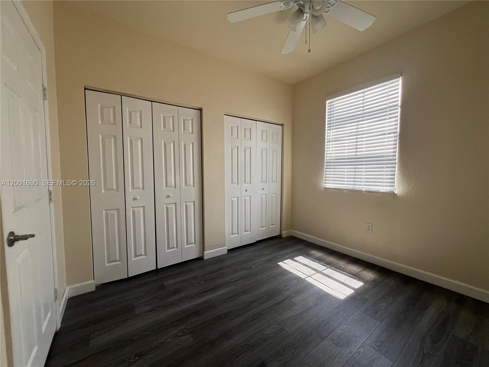 An empty bedroom features dark wood-look flooring, beige walls, two white closets, and a window — 8913 W 35th Way, Hialeah FL 33018, HIALEAH, FL