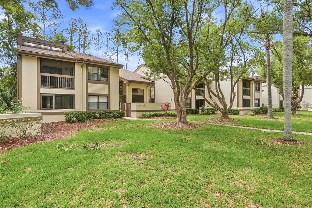A two-story apartment building with tan siding and screened balconies sits behind a grassy lawn — 130 WOODLAKE WYNDE UNIT 130, OLDSMAR, FL