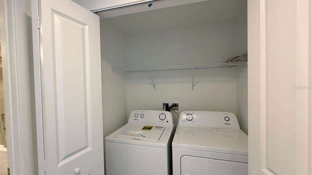 A laundry closet contains a white top-load washer and dryer beneath a wire shelf — 5587 MILLENIA PARK BLVD UNIT 210, ORLANDO, FL