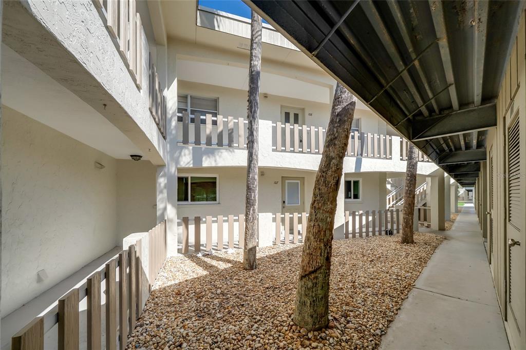 An outdoor walkway leads past a gravel courtyard with two trees between apartment building levels — 6300 FLOTILLA DRIVE UNIT 107, BRADENTON BEACH, FL
