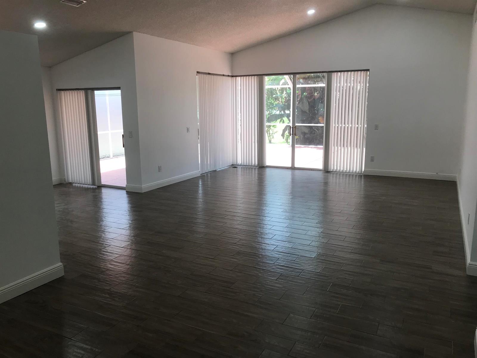 An empty living area with dark wood-look tile flooring, vaulted ceilings, and sliding glass doors — 7296 Via Palomar, Boca Raton FL 33433, BOCA RATON, FL