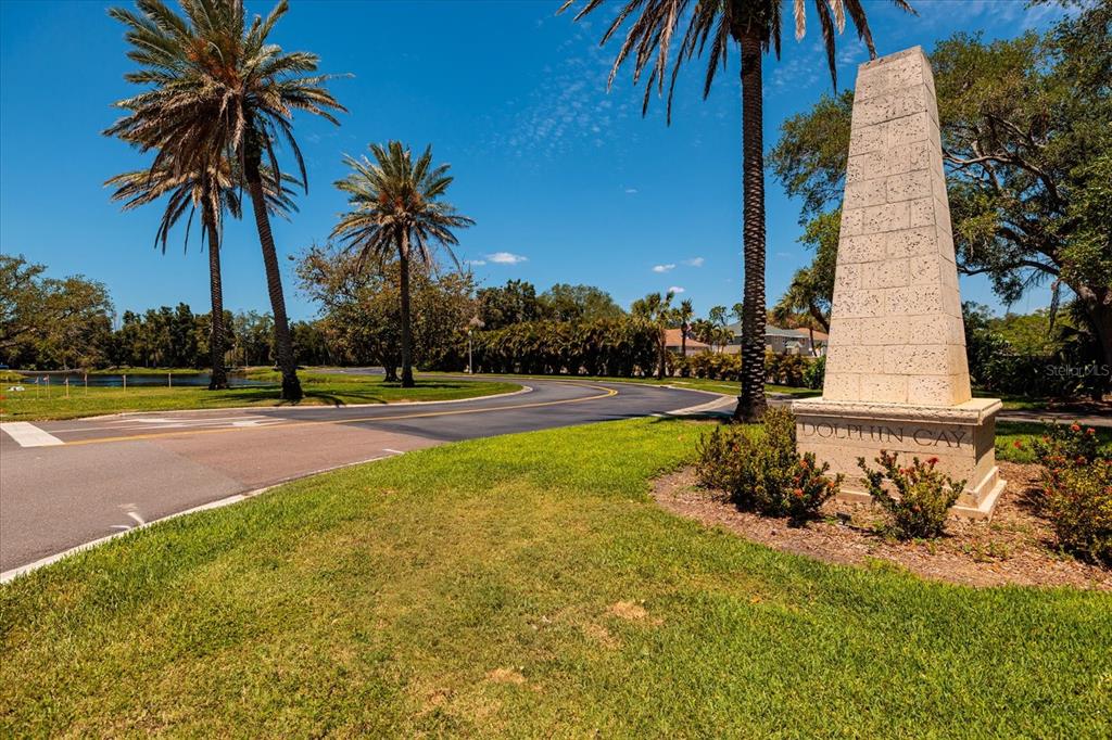 A stone monument with the text Dolphin Cay stands beside a curved road and palm trees — 4801 OSPREY DRIVE S UNIT 207, ST PETERSBURG, FL