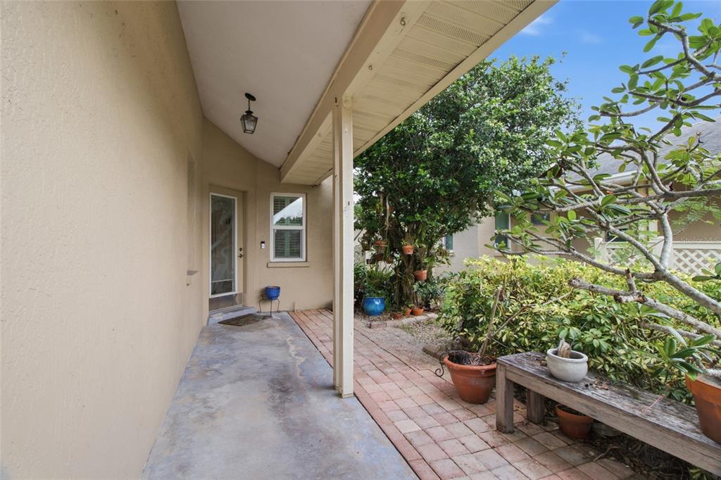 A covered concrete patio leads to a glass-paneled door beside a brick walkway and garden — 308 CHARTLEY COURT N, SARASOTA, FL