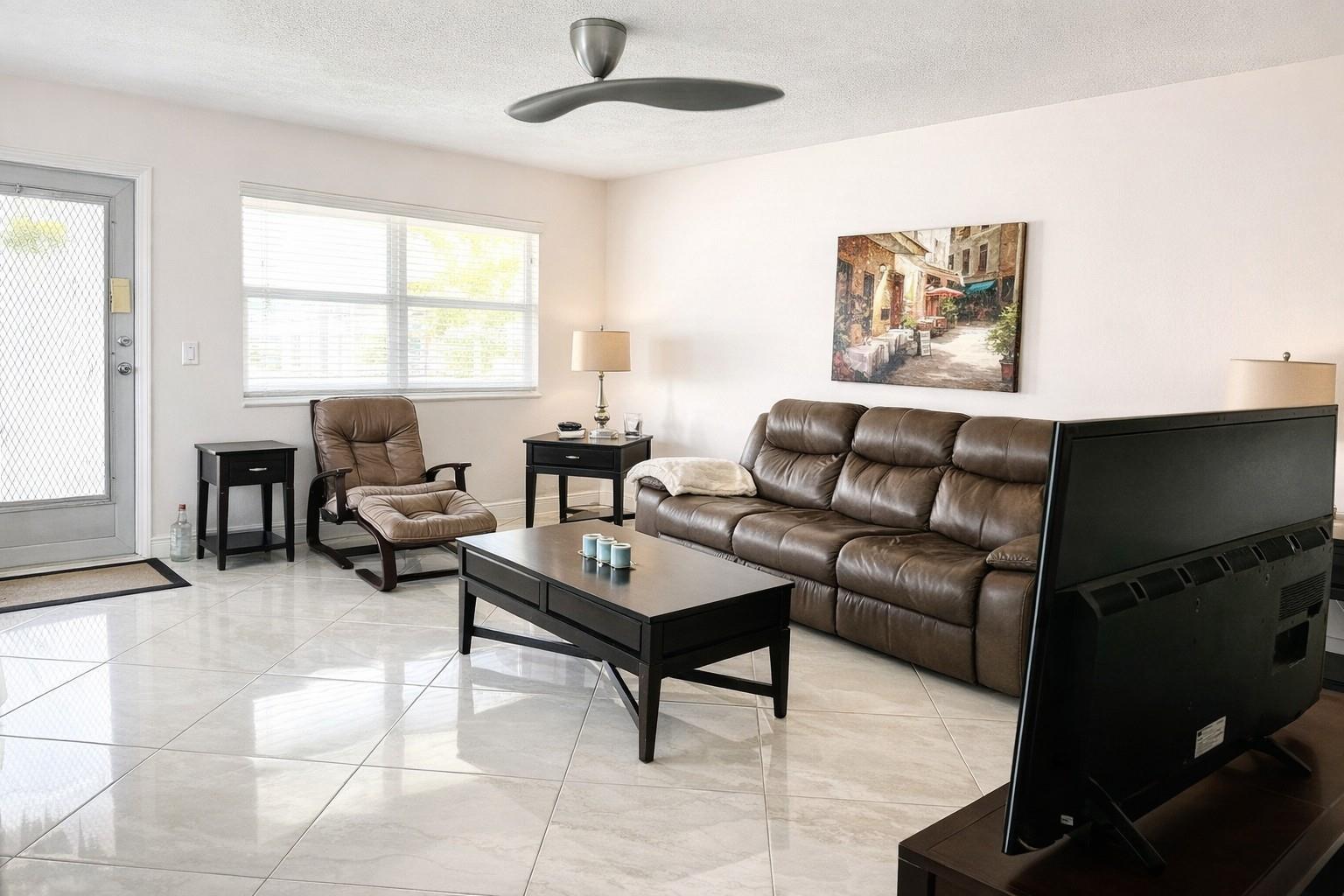 A living room features a brown leather sofa, matching armchair, coffee table, and tiled flooring — 104 E Windsor E # 104, West Palm Beach FL 33417, WEST PALM BEACH, FL