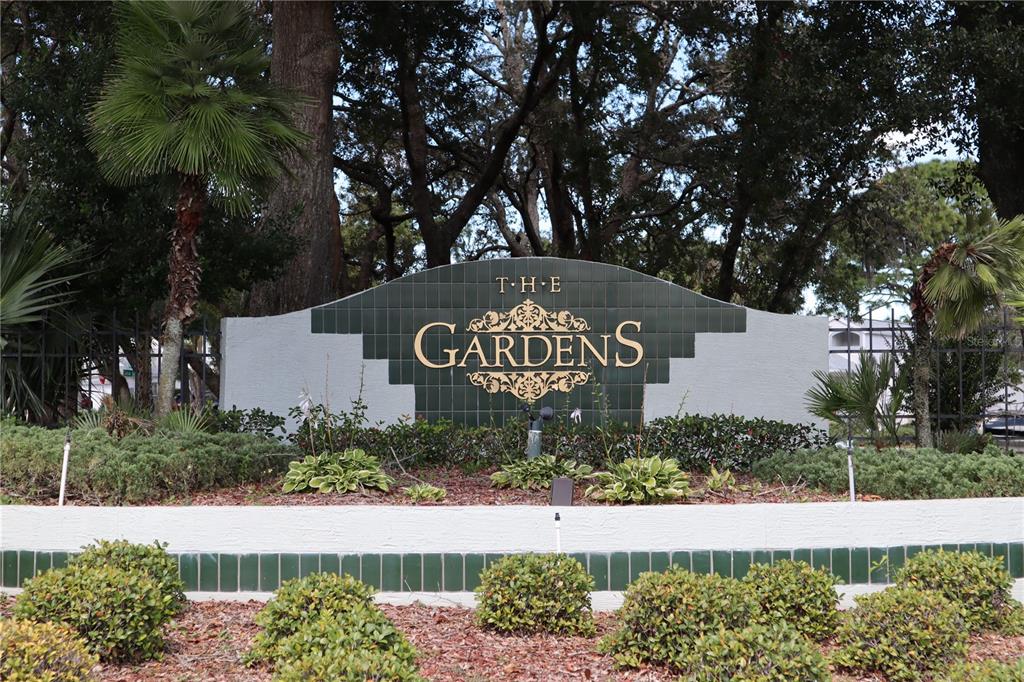 A sign for The Gardens community stands in front of trees and landscaping — 11053 VIA SANTIAGO COURT, SPRING HILL, FL