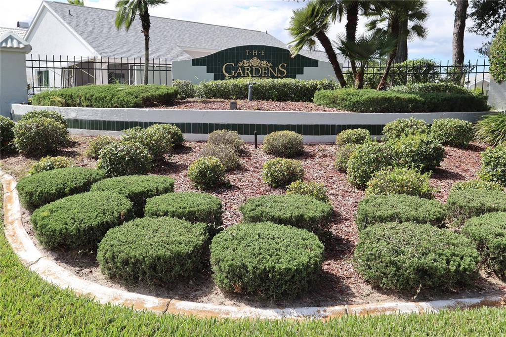A landscaped garden bed features manicured shrubs in front of a sign for The Gardens — 11053 VIA SANTIAGO COURT, SPRING HILL, FL