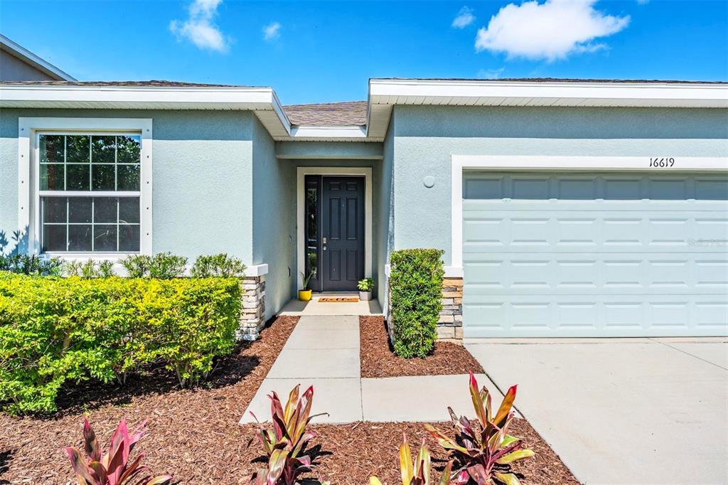 The exterior of a single-story house features a front door, garage, and landscaped front yard — 16619 GOOSE RIBBON PLACE, WIMAUMA, FL
