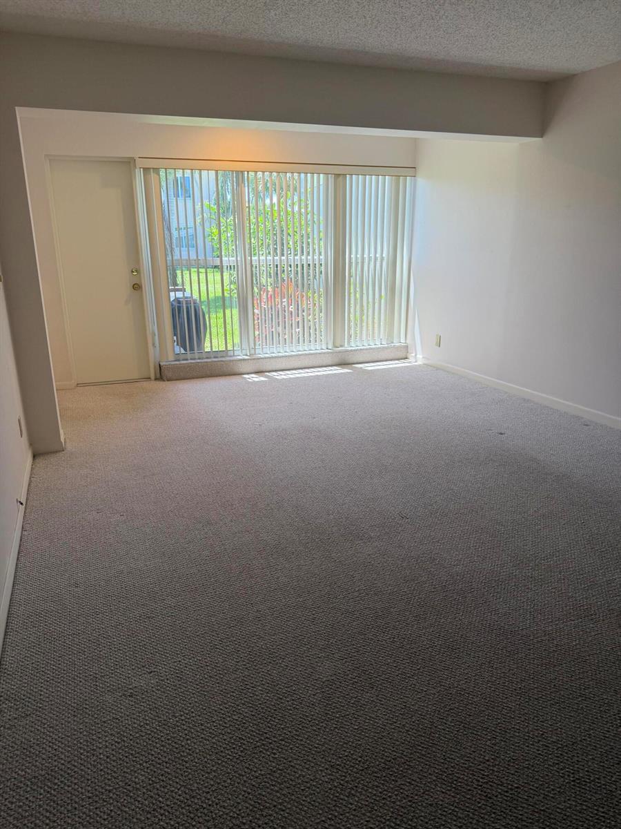 An empty living room features beige carpeting, white walls, and a large sliding glass door — 1360 NW 19th Terrace # 101, Delray Beach FL 33445, DELRAY BEACH, FL