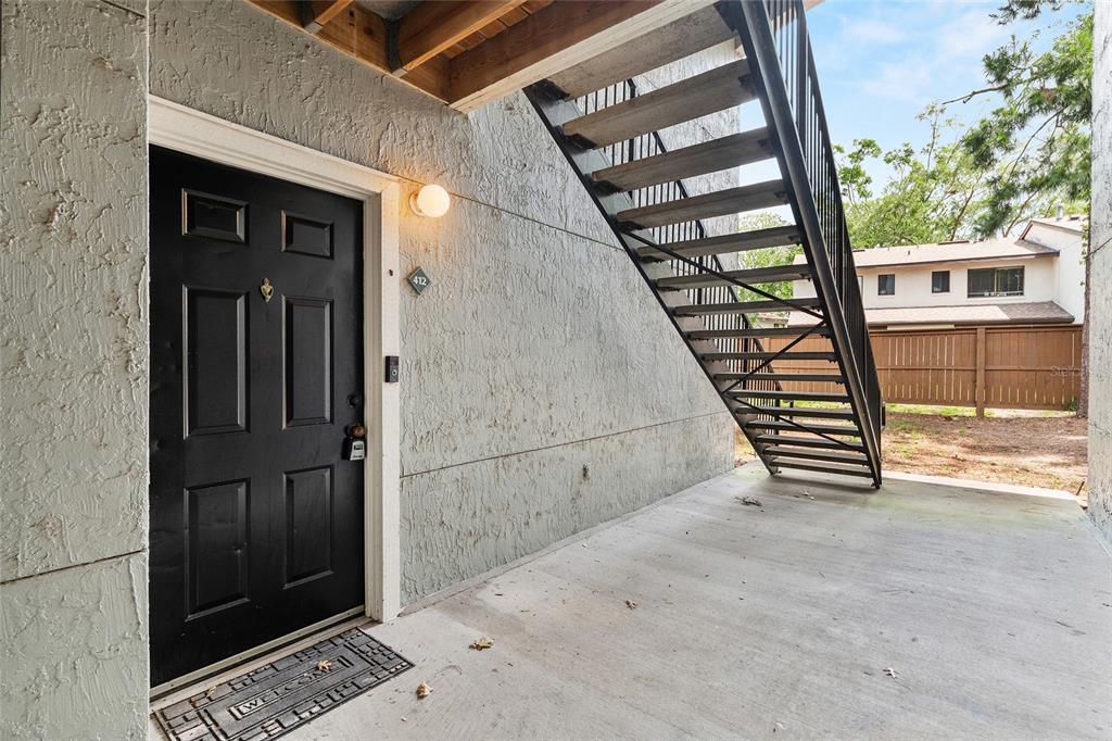 A black front door with a welcome mat sits beneath an outdoor metal staircase — 3705 SW 27TH STREET UNIT 412, GAINESVILLE, FL