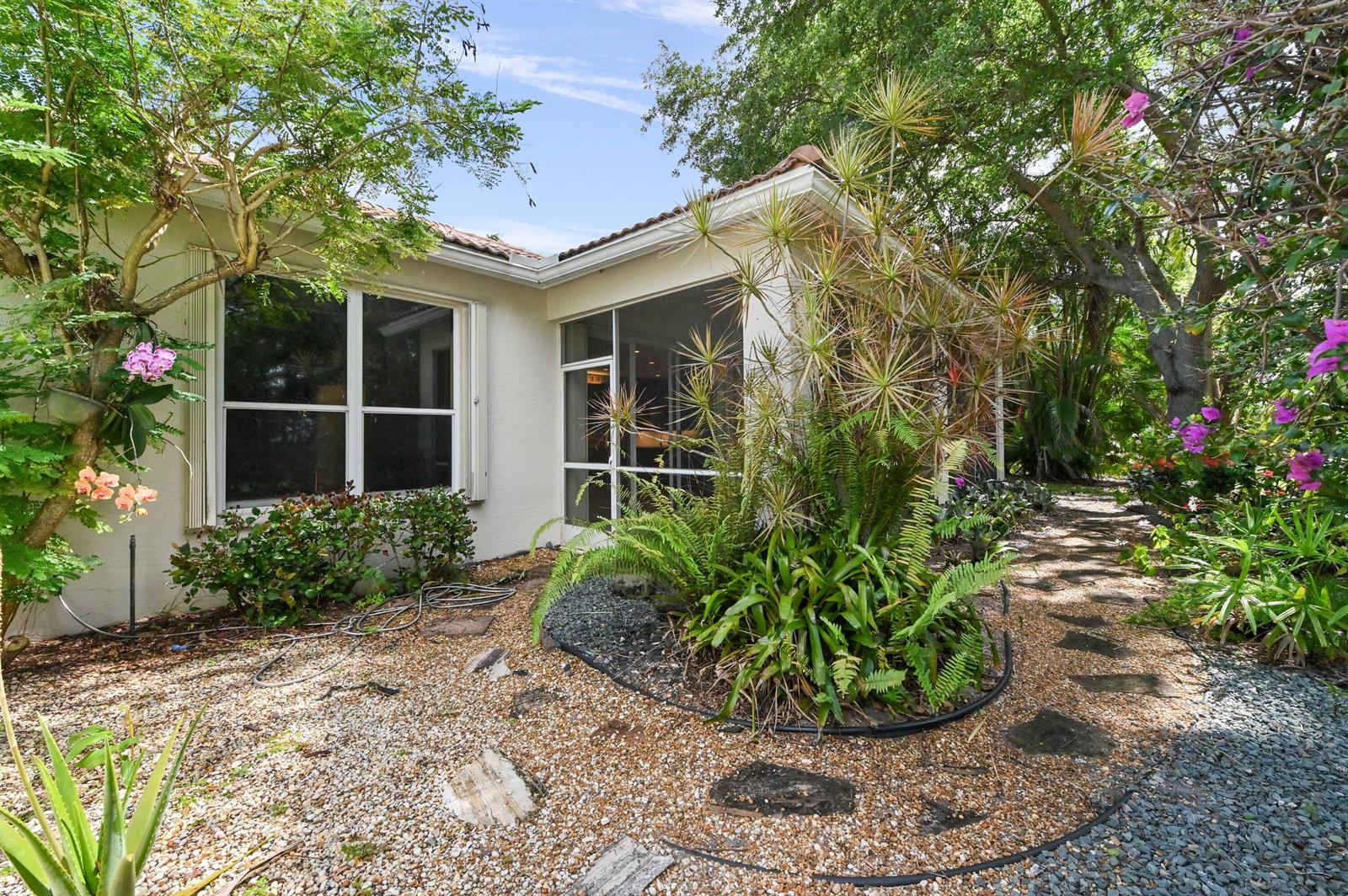 A view of a house exterior with a screened porch, gravel landscaping, and lush greenery — 6439 Pebble Creek Way, Boynton Beach FL 33437, BOYNTON BEACH, FL