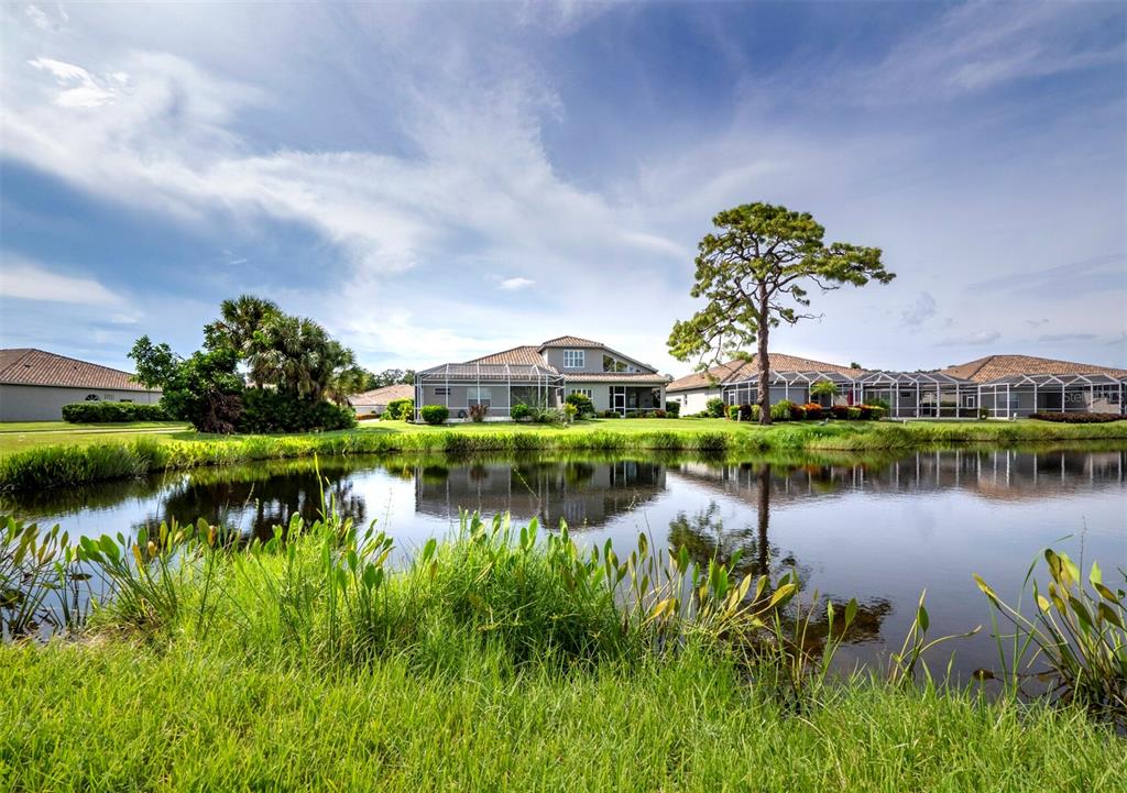 A two-story house with a screened lanai sits behind a pond under a blue sky — 8737 PEBBLE CREEK LANE, SARASOTA, FL