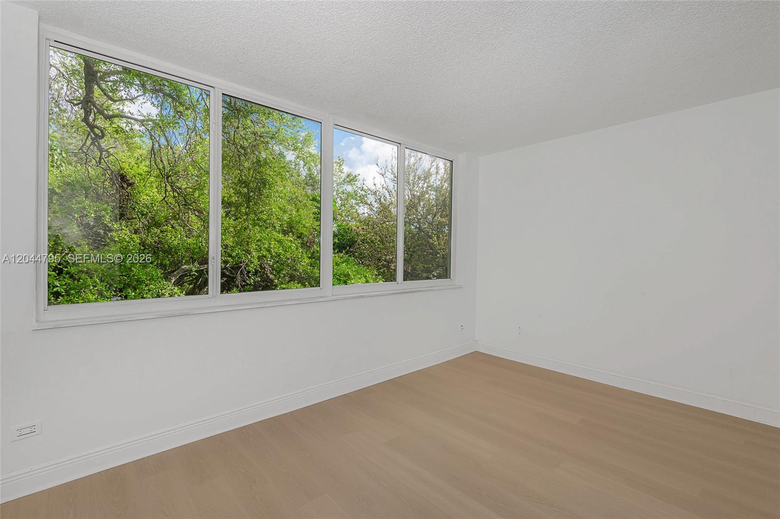An empty room features white walls, light wood flooring, and a large window with greenery — 445 NE 195th St # 326, Miami FL 33179, MIAMI, FL
